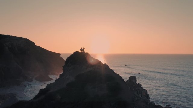 Beautiful aerial shot of female and male surfers standing at the edge of cliff / rock and watching waves at sunrise / sunset. The sun is making a flare. Extreme sport and amazing landscape, nature. Th