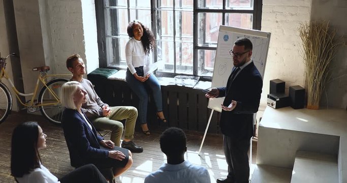 Positive male speaker in eyeglasses presenting marketing research results to smiling mixed race younger and older teammates. Confident trainer coach joking with company staff at workshop in office.