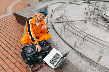Cheerful man in orange clothes and glasses sits with music controller and laptop on the street, plays DJ set and poses for camera with a smile on his face.Musician turns on the music on the DJ console