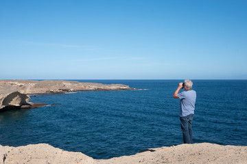 Rear view of elderly man looking with binoculars standing on the cliff - landscape of lava rocks  and blue ocean - free and active retirement concept