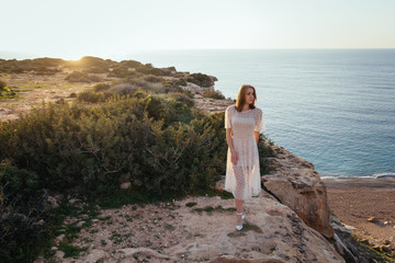 Fototapeta na wymiar A beautiful caucasian young girl in pink dress stay at the edge of cliff and look to the sunset over the sea