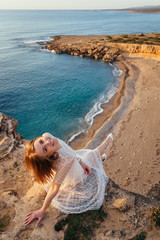 Gorgeous smiling woman in pink dress sits at the edge of cliff and feeling sea breeze at her smiling face. Sunset by the sea.