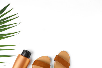 Green palm leaves, brown leather sandals and blank bottle of sun blocking cream as summer symbols, beachwear on white paper textured background with a lot of copy space for text. Top view, flat lay.