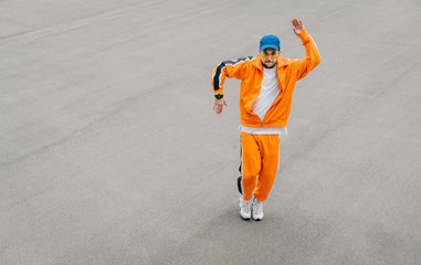 Fashionable young man in stylish casual clothes dancing modern hip hop dancing on a background of gray asphalt, wearing an orange tracksuit. Dancer shows hip hop performance isolated on asphalt.