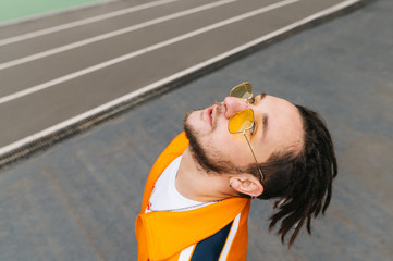 Portrait of a stylish young man with dreadlocks and sunglasses, wearing an orange tracksuit,...