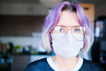 Worried woman in a medical mask and glasses looks to the camera inside her house. 