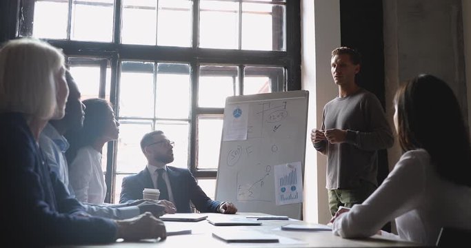 Focused young male speaker giving presentation on flipchart to interested different ages diverse business people. Smart presenter coach trainer pointing on whiteboard, explaining strategy at office.