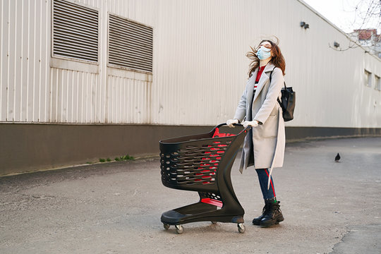 Young Woman With Beautiful Blue Eyes Wearing Protection Face Mask Against Coronavirus COVID-2019, Novel Coronavirus 2019-nCoV Pushing A Shopping Cart On Supermarket Department Store Wall Background