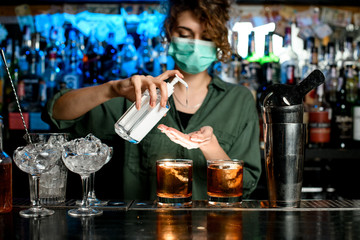 Woman bartender in medical mask treats her hands with disinfector