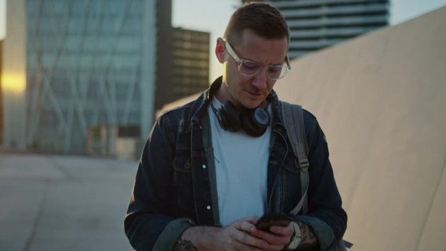 Slow Motion of Attractive Man Photographing with his Smartphone While Walking. Male Person with Glasses and Headphones Using Phone to Take Pictures While Walking. Backward Tracking Cinematic Shot