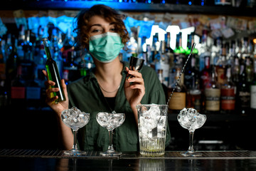 bartender girl in medical mask holding bottle of green liquid in one hand and beaker in other hand preparing to make mixture.