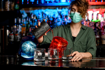 bartender girl in medical mask carefully pour drink from steel glass into glasses with ice.