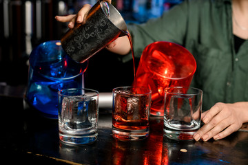 Close-up bartender girl in medical mask carefully pour drink from steel glass into glasses with ice.
