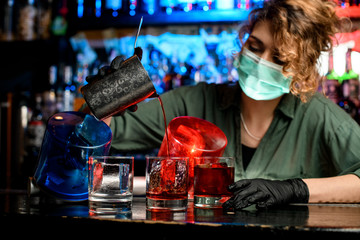 Young handsome bartender girl in medical mask and black gloves accurate pour drink from steel glass into glasses with ice.