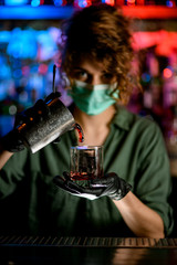 Woman barman in medical mask and black gloves holds glass and energetically pours drink into it.