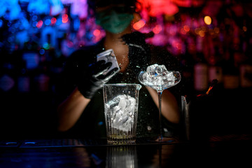 Close-up. Woman barman in medical mask and black gloves with special device accurate breaks pieces of ice.