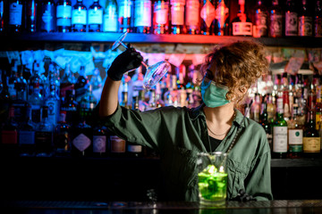 young girl at bar counter in medical mask and black gloves holds glass.