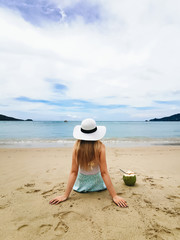 Fototapeta na wymiar Young attractive girl in a hat on the shore of the blue sea sitting back with a coconut, Thailand