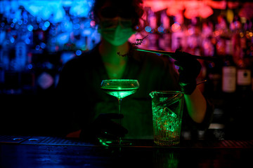 young girl at bar counter in medical mask and black gloves holds glass with green cocktail and decorates it by flowering branch.