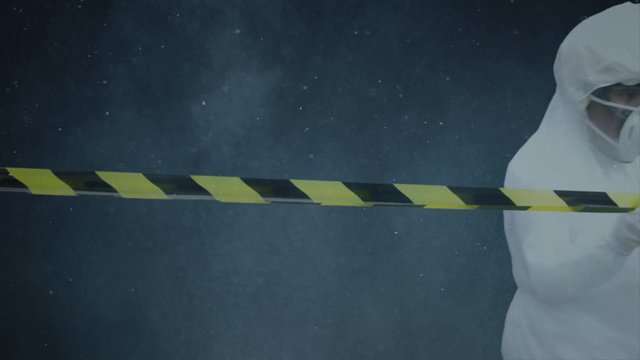Woman with bacteriological protection suit rolling a striped warning tape in the night. Copy space for text or other content
