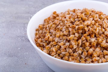 Cooked buckwheat in white ceramic bowl on grey background. Close up, horizontal format.