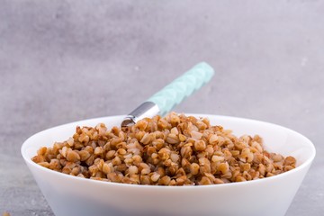Cooked buckwheat in white ceramic bowl and with spoon on grey background. Close up, space for text, horizontal format.