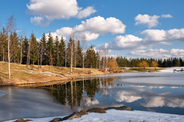 Fototapeta na wymiar Spring landscape. Forest lake with reflection of clouds and trees growing along the shore, partially covered with ice and blue sky with clouds, at the beginning of a spring day.