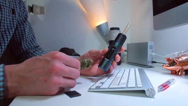 An office worker sitting at a table in front of a computer holds a vaporizer and marijuana before use. The concept of psychological relaxation of man.