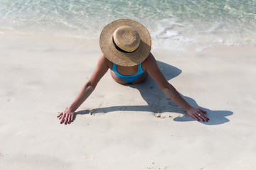 Fototapeta na wymiar Rear view of young woman in bikini and straw hat sitting on beach