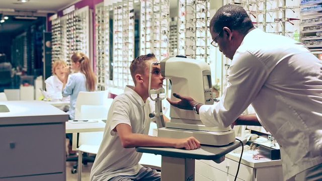 Teenage boy receiving eye examination on modern refractometer in optical store with professional African-American ophthalmologist 