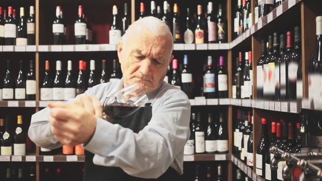 Confident elderly male winemaker holding glass of red wine, checking it in wine store