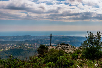 Fototapeta na wymiar A metal cross installed on top of a cliff of the Baou des Noirs near Vence, France in the Alps mountains with the landscape and Mediterranean Sea in the background (Cote d'Azur / Provence)