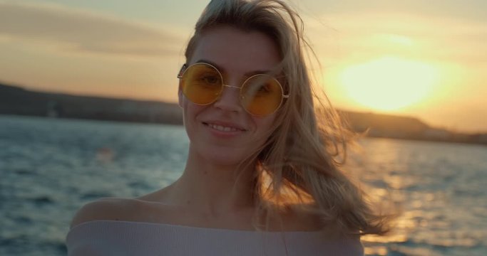 Portrait happy blonde woman smiling in yellow sunglasses runs hand over face standing seashore looking camera background sea city port orange sunset sunny day, lens flare. Hair fluttering wind