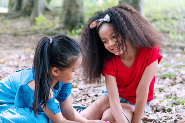 Cheerful two kid girl holding plasticine looking each other while sitting together in the park. Mixed race schoolchild with plasticine in her hands while playing with asian friend. Friendship concept.