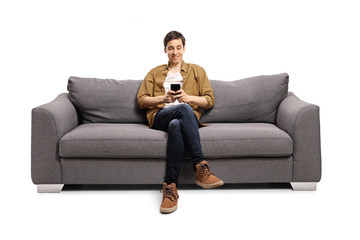 Happy young man sitting on a gray sofa and typing on a mobile phone