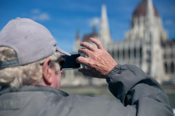 Old elderly pensioner tourist makes a photo of the sights of the parliament building in Budapest