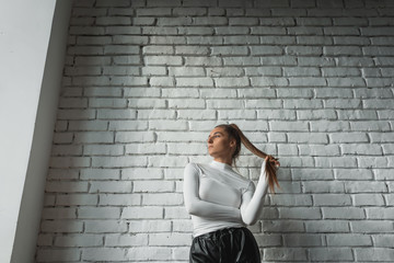 Fashionable brunette girl in a white sweater and black leather pants posing on an white bricks...