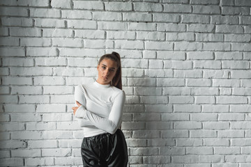 Fashionable brunette girl in a white sweater and black leather pants posing on an white bricks background.