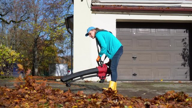 Hired female worker blowing leaves from garage entrance in autumn