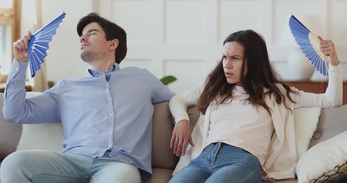 Unhappy frustrated couple suffering from heat, bad air conditioning at home waving fans sitting on sofa. Overheated young man and woman sweating feeling hot and dehydrated in summer swelter concept.
