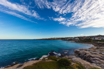 The rocky shoreline around Bondi beach, Sydney, Australia. Waves splashing at the rocks in the water. White clouds on blue sky. The cliffs beside the sandy beach. A paradise for surfers