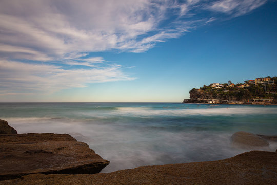 The rocky shoreline around Bondi beach, Sydney, Australia. Waves splashing at the rocks in the water. White clouds on blue sky. The cliffs beside the sandy beach. A paradise for surfers 