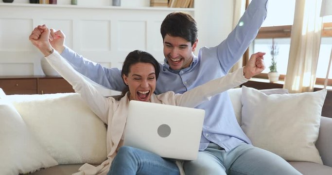 Overjoyed young family couple winners sport fans watching game on laptop celebrating victory goal sit on sofa. Excited man and woman shoppers winning online shopping voucher feeling amazed together.