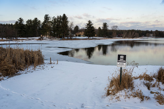 A metal sign marking a no-boat launch area, stands in the ground on the shore of a half frozen lake covered in ice and snow.
