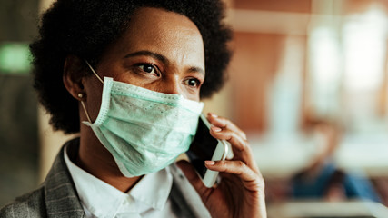 Black businesswoman wearing face mask and communicating on cell phone in public area.