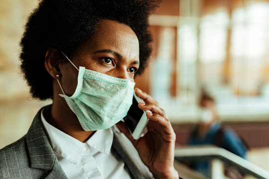 Black Businesswoman Communicating On Mobile Phone While Wearing Protective Mask At Airport Corridor.