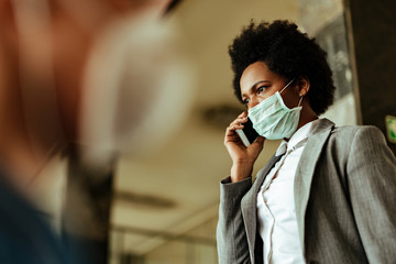 African American businesswoman communicating on smart phone while wearing face mask at public corridor.
