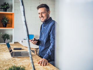 happy smiling remote online working man with laptop, mobile phone and notebook in casual outfit standing up in front of a work desk in an coworking office