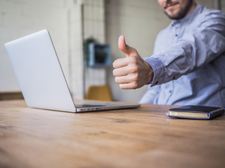 thump up next to laptop and notebook of a happy smiling remote online working man typing and in...