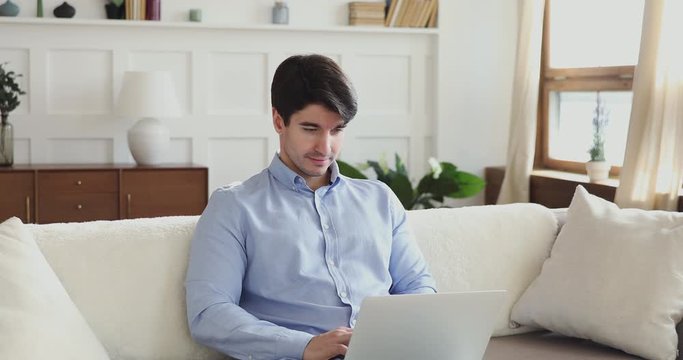 Millennial man using laptop sitting on couch in living room. Young guy browsing internet, surfing web, reading online news, typing on notebook computer modern technology device relaxing at home.